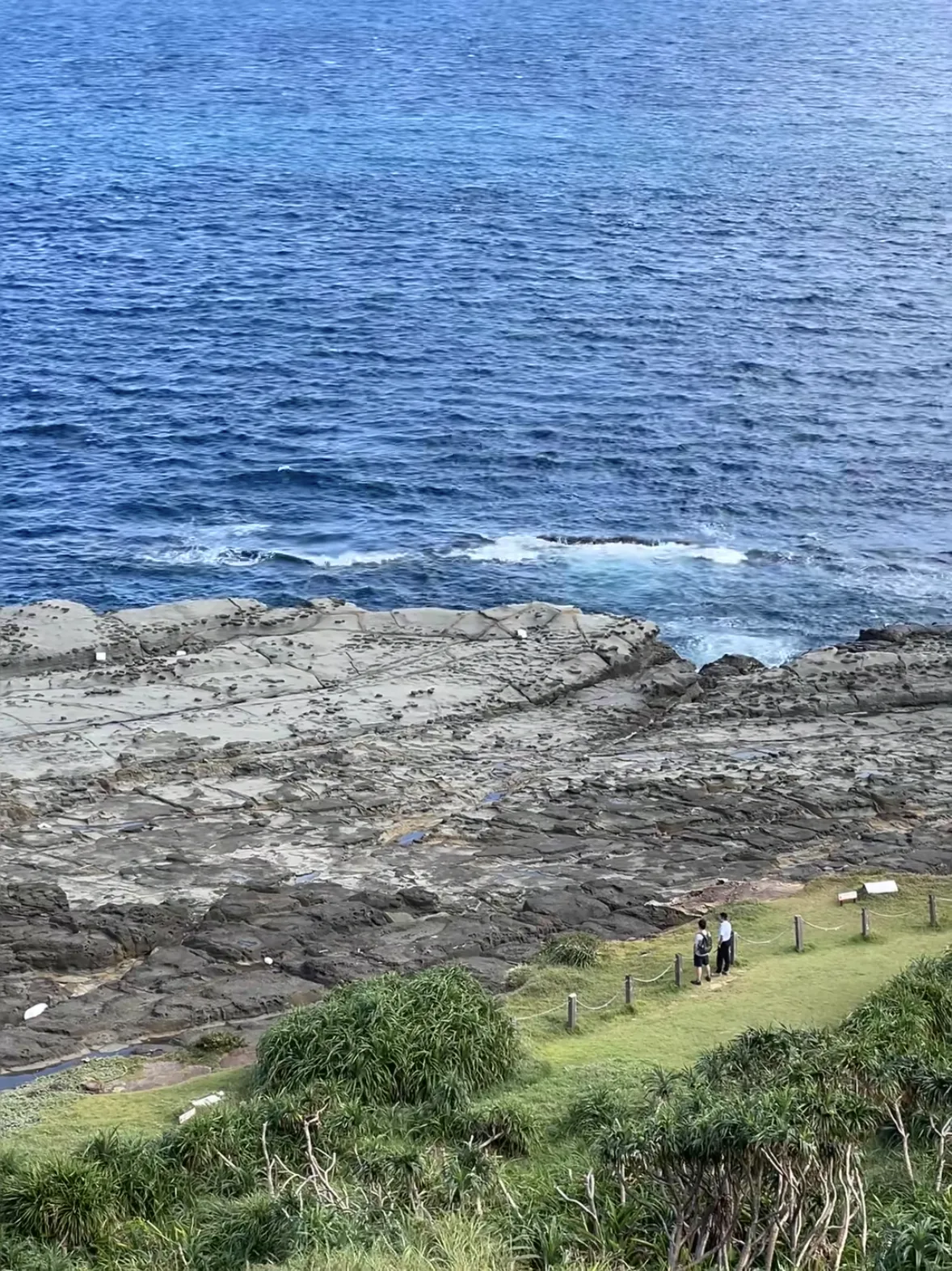 Two people standing on a wide sandstone shelf where green headland meets deep blue sea on Taiwan's northeast coast
