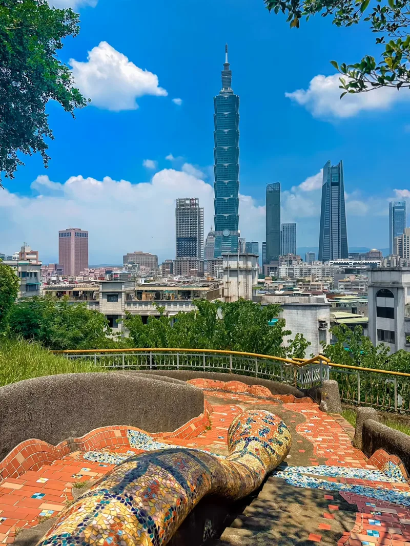 Taipei 101 rising over the city under blue sky, seen from a hillside park with a mosaic-tiled sculpture in the foreground